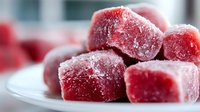 Frozen red fruit cubes covered in frost crystals on white plate, macro food photography showing texture and cold temperature details in soft natural light.