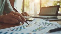 Businessman analyzing financial charts and graphs with pen, reviewing data analytics and business performance metrics on desk with laptop in bright office environment.