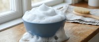 White foamy shaving cream in blue ceramic bowl on wooden surface near window with natural light and grooming accessories for men's skincare routine.