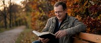 Middle-aged Caucasian man with glasses reading book on park bench during autumn season with colorful fall foliage background for lifestyle content.