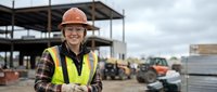 Smiling female construction worker in hard hat and safety vest at building site with heavy machinery and concrete structure in background.