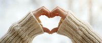 Hands in a cozy knit sweater forming a heart shape against a soft winter background, symbolizing love, warmth, and care.