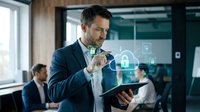 Businessman in dark suit holding tablet with glowing digital security icons, padlock and data protection symbols displayed on screen in modern office environment with colleagues in background.