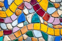 Colorful mosaic pattern made with irregular stone and ceramic pieces in vibrant yellow, blue, red, pink, and turquoise creating abstract wavy lines on textured surface.
