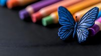 Vibrant blue butterfly resting on colorful art supplies against dark background, creating artistic composition with selective focus and shallow depth of field.