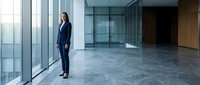 Young professional woman with long dark hair, South Asian ethnicity, wearing formal navy business suit standing in modern empty office space with floor-to-ceiling windows and concrete flooring.