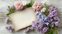 Blank vintage paper card decorated with fresh spring flowers - pink carnations and purple lilac blooms on rustic wooden background. Romantic floral layout.
