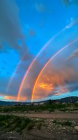 Spectacular double rainbow arches across dramatic sunset sky with glowing orange clouds and mountain silhouettes, rural landscape below creates inspiring natural phenomenon.
