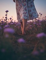 Barefoot woman in floral dress standing among purple wildflowers at sunset, creating dreamy atmospheric mood with soft focus foreground flowers and golden light.