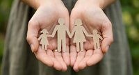 Hands holding wooden family figures representing protection, care and unity in family insurance and social support concepts.