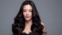 Young Asian woman with long wavy black hair and natural makeup against gray background, professional beauty portrait showcasing healthy shiny hairstyle and flawless skin.