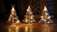 Illuminated glass Christmas trees with warm fairy lights creating festive holiday ambiance on wooden surface against decorative background.