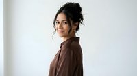Young Hispanic woman with curly dark hair in brown blazer smiling over shoulder in bright modern office space for professional business portraits.