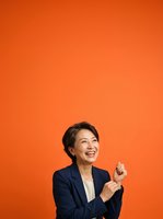 Mature Asian woman in dark blazer and white shirt smiling and gesturing upward against vibrant orange background, suitable for business, corporate, leadership, and professional success concepts.