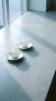 Two white ceramic cups with saucers sit on a concrete table near bright windows, casting soft shadows in natural daylight. Minimalist cafe setting.