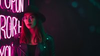Young woman with long hair in leather jacket standing near neon pink sign in dark urban nighttime setting with colorful lighting for lifestyle content.