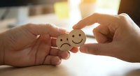 Hands exchanging wooden emotion tokens showing sad and happy faces in warm sunlight. Mental health awareness and emotional support concept for wellness campaigns.