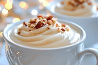 Creamy whipped coffee drink topped with crushed pecans in white ceramic cup against soft bokeh background, close-up view showing luxurious texture and garnish.