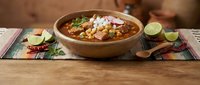 Traditional Mexican pozole soup with hominy corn and meat served in rustic wooden bowl with lime wedges and colorful textile placemat on wooden table.