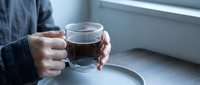 Person holding clear glass mug with hot tea or coffee by window, cozy morning routine, warm beverage, relaxation and comfort at home.