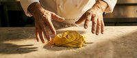 Professional chef hands kneading fresh pasta dough on floured wooden surface in warm kitchen lighting for culinary cooking preparation.