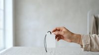 Caucasian woman's hand holding eyeglasses with black frames against neutral interior background, suitable for optometry, vision care, eyewear retail, and healthcare marketing materials.
