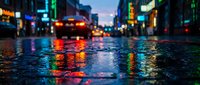 Wet urban street at dusk reflects neon lights from storefronts and vehicles, creating vibrant bokeh effects. Suitable for backgrounds, city themes, and nightlife concepts.