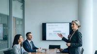 Mature female professional with gray hair, wearing business attire, presenting to two colleagues in modern office conference room with wall-mounted screen. Corporate training, meeting, leadership concept.
