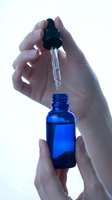 Hands holding blue glass dropper bottle with pipette for skincare serum or essential oil application on white background.