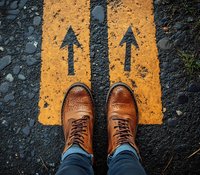 Brown leather boots standing at yellow arrows painted on asphalt road, personal perspective view showing choice, direction and decision making concept.