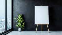 Empty white canvas on wooden easel against dark wall near window with urban view and decorative plant in modern minimalist interior space.
