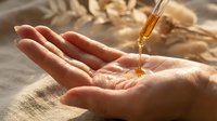 Golden oil drops from pipette into cupped hands with wheat ears in background. Natural skincare, aromatherapy, wellness treatment concept.