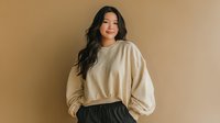 Young Asian woman with long dark hair wearing cream oversized sweatshirt and black skirt against neutral beige background, casual fashion portrait.