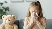 Young Caucasian woman blowing nose in tissue while sitting on couch with teddy bear, showing cold and flu symptoms during illness at home. Healthcare concept.