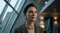 Middle-aged woman with dark hair and fair complexion, wearing a gray cardigan and pearl necklace, gazing thoughtfully in modern office interior with large windows and contemporary lighting.