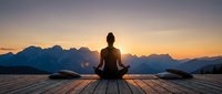 Woman practicing yoga meditation at sunrise on wooden deck overlooking mountain landscape. Peaceful morning wellness routine with dramatic golden sky backdrop.