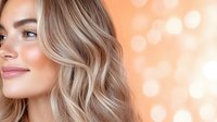 Young Caucasian woman with wavy blonde hair and natural makeup smiling against blurred warm bokeh background, side view portrait showing radiant glowing skin.