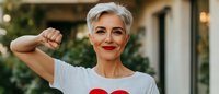 Confident mature woman with short gray hair and red lipstick wearing white t-shirt with heart print smiling outdoors on blurred background. Lifestyle portrait.
