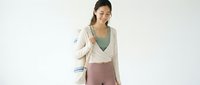 Young woman with dark hair and olive complexion wearing casual layered outfit of cream cardigan and mauve pants, smiling at camera against white background for lifestyle and fashion content.