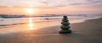 Zen stone stack on sandy beach at sunset with gentle waves and golden sky reflection creating peaceful meditation atmosphere for wellness and mindfulness practices.