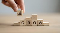 Hand placing wooden block with growth chart icon on top of stair-step arrangement of blocks spelling GROW, symbolizing business development and success.