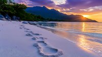 Footprints along pristine white sandy beach at dramatic purple and orange sunset, waves washing shore with mountain silhouettes and glowing sun on horizon.