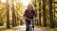 Happy senior Caucasian woman riding bicycle in autumn park with golden sunlight filtering through trees, enjoying active outdoor lifestyle and exercise.