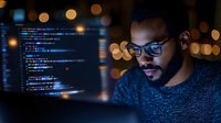 Young African American software developer coding at night with colorful bokeh lights and programming code on computer screen in modern office workspace.