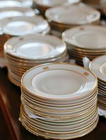 Stacks of elegant white porcelain dinner plates with gold trim arranged on wooden surface, showcasing fine dining tableware for restaurant and catering settings.