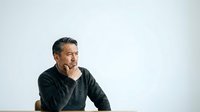 Middle-aged man with dark hair and olive complexion wearing dark sweater, hand on chin in thoughtful pose against white background for business and editorial use.