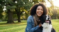 Happy young African American woman laughing while hugging her Border Collie dog in sunny park during golden hour autumn day outdoors.