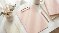 Elegant pink clipboard with rose gold pen on white marble desk surrounded by dried flowers, notebooks and coffee mug for feminine office workspace styling.