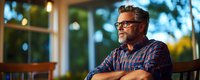 Mature bearded man with glasses in plaid shirt sitting thoughtfully on outdoor patio during golden hour sunset with warm bokeh lighting and home exterior.