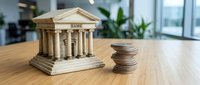 Wooden bank building model with classical columns next to stack of coins on office desk representing financial services and banking concepts.